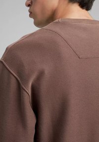 Suéter marrón de punto acanalado con un acabado texturizado y detalles de costuras visibles en los hombros. La vista se centra en la parte trasera de la prenda.