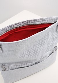 Stříbrná kabelka s textura a geometrickým vzorem, červenou podšívkou, zipovým uzávěrem a stříbrnými kovovými akcenty.