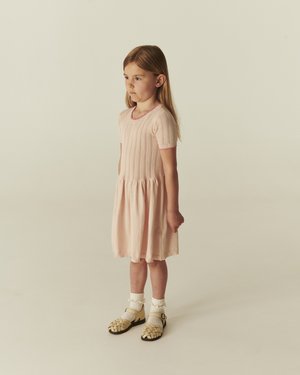 Jeune fille aux longs cheveux portant une robe rose pâle à manches courtes, des chaussettes à volants et des sandales tissées beige, debout devant un fond uni.