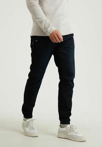 Granatowe tapered spodnie z denimu z wzmocnionymi łatami na kolanach, zestawione z białą bluzką z długim rękawem i białymi sneakersami.