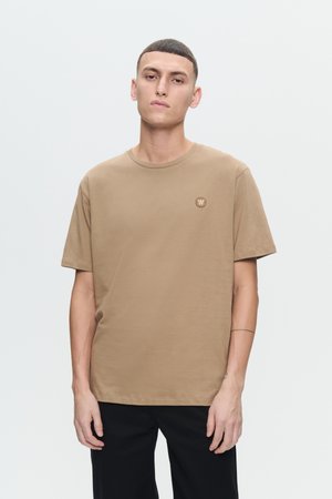 Young man with short dark hair wearing a plain brown t-shirt with a small circular logo on the chest and black pants, standing against white background.
