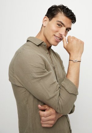 Joven con cabello oscuro que lleva una camisa marrón claro abotonada, sonriente y mostrando una pulsera plateada con nudos en su muñeca levantada.