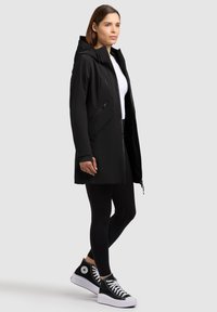 Donna in piedi con cappotto nero con cappuccio, camicia bianca, leggings neri e sneakers alte nere su uno sfondo chiaro e uniforme.