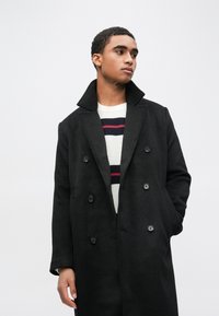 Sort dobbeltradet frakke med store revers, fremtrædende tekstureret uldstof, rummelig pasform og matchende knapper. Indvendig sweater har dristige striber.