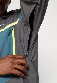 Casaco leve cinza com realces em teal e amarelo fluorescente, apresentando um bolso lateral com fecho e um toque elegante e suave.