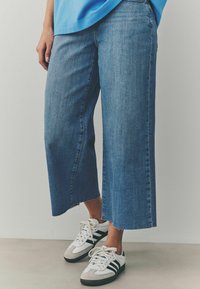 Jeans de mezclilla acortados y de pierna ancha en azul medio. Presentan un dobladillo deshilachado, un corte recto y una sutil textura. Combinados con zapatillas negras y blancas.