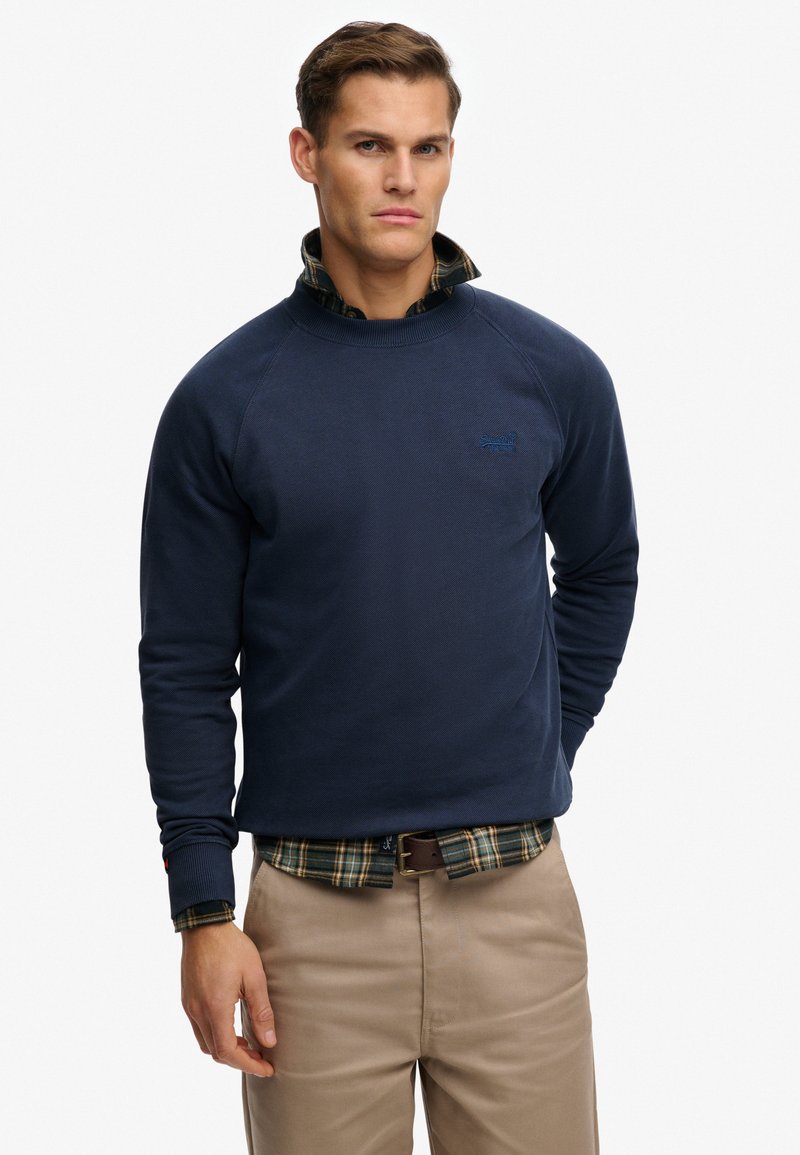 Felpa a girocollo blu navy con maniche raglan, tessuto strutturato e piccolo logo; indossata sopra una camicia a quadri verde e pantaloni beige.