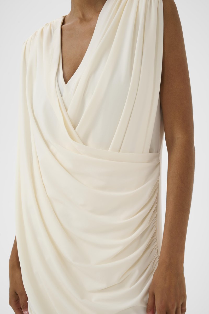 Robe sans manches crème drapée avec un décolleté en V profond, détails de tissu froncés sur le côté, portée par une personne à la peau brune sur un fond uni.