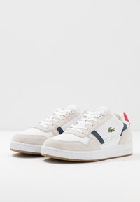 Baskets blanches et beiges avec tige en daim et cuir, bande marine, accent rouge au talon et semelle en caoutchouc texturée. Logo Lacoste sur le côté.