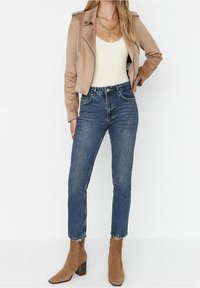 Chaqueta corta de ante marrón claro, top ajustado color crema, jeans de tiro alto azul oscuro y botines color beige con tacón cuadrado.