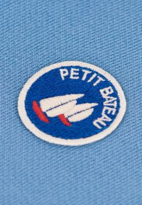Vyšívané kruhové logo na svetlomodrom látkovom podklade, so modrým pozadím, bielou plachetnicou, červenými akcentmi a textom "PETIT BATEAU".