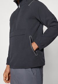 Zwarte kwart-rits jas met een hoge kraag, met grijze accenten, een voorzak en elastische manchetten. Gemaakt van lichtgewicht, gestructureerde stof.