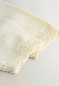 Helevalge koorivärvi kudumiga kampsuni detail, millel on soonikkoes varrukaservad, esitades tekstuurse kanga ja pehme kootud mustri, volditud neutraalsel taustal.