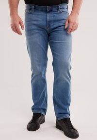 Mees, kes kannab helesiniseid teksapükse ja musti tossusid, seisab käed vabal asendil keha kõrval ühevärvilise tausta ees.