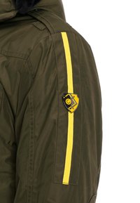 Grüne gepolsterte Jacke mit gelbem Seitenstreifen, ausgestattet mit einem schildförmigen Logo auf dem Oberarm. Das Material wirkt glatt und strapazierfähig.