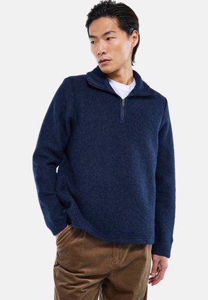 Personne portant un pull à demi-fermeture éclair bleu marine sur une chemise blanche et un pantalon marron, posant avec une main dans la poche, regardant vers la gauche.