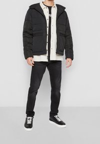 Svart puffjacka med huva, med horisontell sömnad och två sidofickor. Bärs över en ljus skjorta och svarta jeans med sneakers.