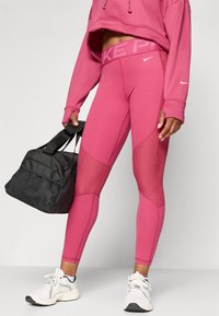Różowe legginsy z siateczkowymi wstawkami, wysoki pas z logo "NIKE", połączone z czarną torbą sportową. Białe buty sportowe dopełniają całość.