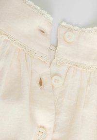 Gros plan sur un tissu crème avec une délicate bordure en dentelle et trois boutons blancs assortis à leurs boutonnières.