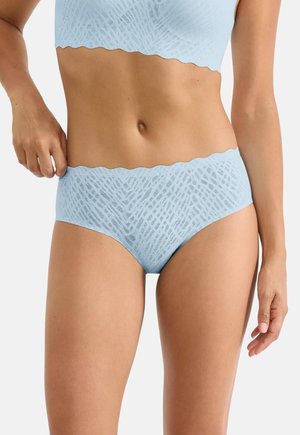 Donna che indossa biancheria intima in pizzo senza cuciture azzurro chiaro e reggiseno coordinato, tenendo in mano la vita della biancheria su sfondo bianco neutro.