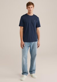 Mlad moški stoji v mornarsko modri majici s kratkimi rokavi, svetlih modrih kavbojkah in belih športnih copatih na enotnem bež ozadju.