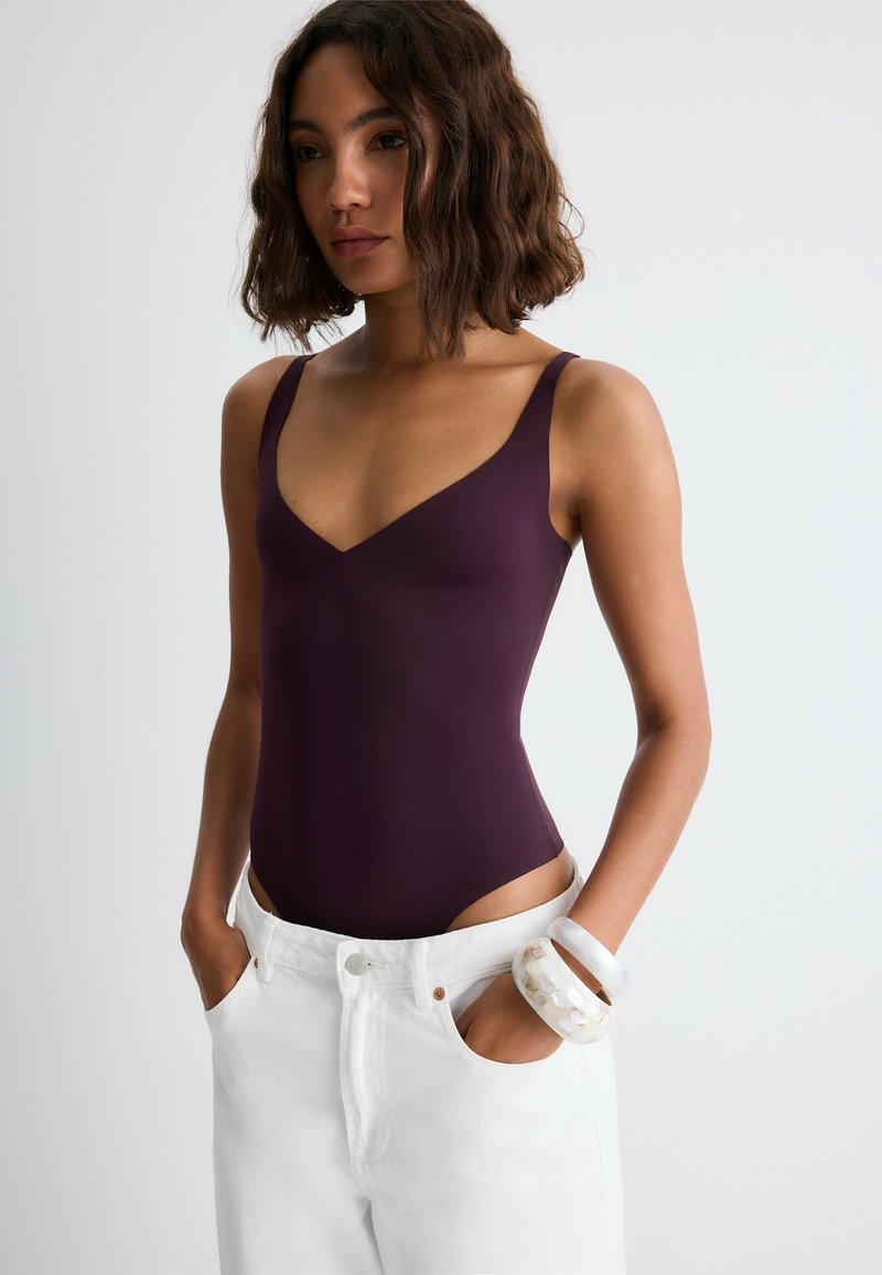Femme aux cheveux ondulés jusqu’aux épaules porte un body sans manches violet foncé, un pantalon blanc et deux bracelets épais, les mains dans les poches.