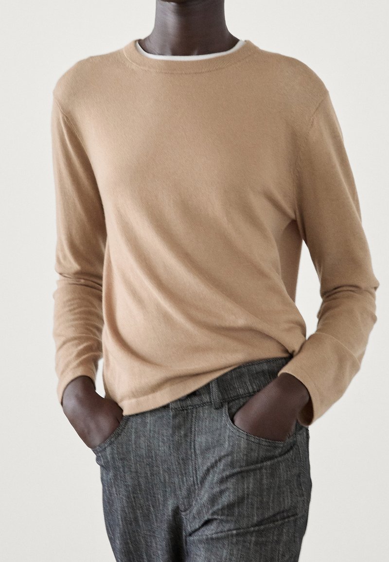 Homme portant un pull beige à manches longues et un pantalon gris texturé, les mains dans les poches, debout devant un fond clair uni.