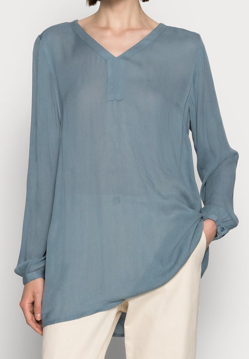 Personne portant un chemisier bleu ample à manches longues avec un col en V et un pantalon crème, une main dans la poche, sur un fond uni.