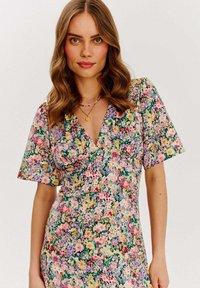 Květinové šaty s různými barevnými květinami na zeleném pozadí, krátkými rukávy, hlubokým výstřihem do V a přiléhavým pasem.
