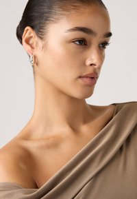 Τοπ σε χρώμα επιδερμίδας που πέφτει πάνω στον ώμο, μαλακό ύφασμα με λεία υφή· το μοντέλο φοράει ασημένιους κρίκους· το δέρμα είναι ελαφρώς φωτισμένο.