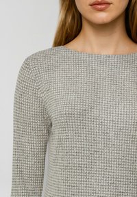 Vrouw in een lichtgrijs en witte geblokte, gestructureerde blouse met lange mouwen en een ronde halslijn.