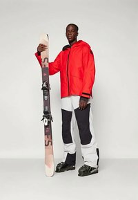 Piros vízálló dzseki, fekete kiegészítésekkel, magas gallérral; fehér és fekete sínadrág; síléceket tart, amikhez kötések tartoznak; texturált sícipő.