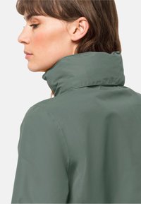 Veste verte imperméable avec un col haut et une texture lisse, dévoilant les épaules et une partie du cou. Design minimaliste sans quincaillerie visible.