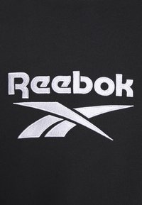 Reebok-logotyp broderad med vit tråd på svart tyg, med fet text och en abstrakt vektorsymbol under varumärket.