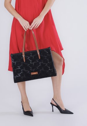 Mujer con vestido rojo sosteniendo un bolso tote de encaje negro con asas marrones, usando zapatos de tacón alto tipo slingback negros.