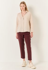 Camicia button-up in lino beige a maniche lunghe, abbinata a pantaloni in lino bordeaux. Il modello indossa sneakers chiare, in piedi su uno sfondo neutro.