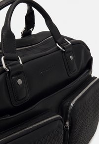 Sac en cuir noir avec une texture tissée, des accessoires en argent, des fermetures éclair doubles et des poignées robustes pour un transport pratique.