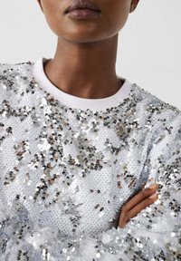 Close-up van een persoon die een zilverkleurige top bedekt met pailletten draagt met gekruiste armen, waarbij de halslijn en een deel van het gezicht zichtbaar zijn.