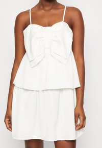 La robe blanche présente des bretelles fines ajustables, un design en couches et un nœud proéminent sur la poitrine avec une texture de tissu lisse.