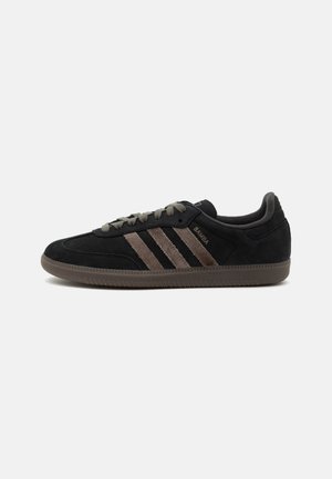Baskets noires Adidas Samba avec des bandes en suède marron, des lacets assortis et une semelle en caoutchouc marron, vues de côté sur fond blanc.