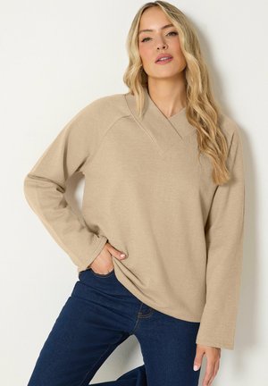 Blonde Frau trägt ein beige langärmeliges Oberteil mit weitem Kragen und dunkelblaue Jeans, posiert mit einer Hand in der Tasche vor einem einfarbigen Hintergrund.