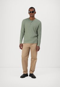 Zöld, hosszú ujjú henley ing egy textúrált felülettel, khaki nadrág és fekete cipő. A modellen napszemüveg van, egy sima háttér előtt áll.