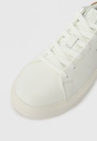 Λευκά δερμάτινα αθλητικά παπούτσια με παχιά λευκή κορδέλα δεμένη σε φιόγκο, στρογγυλή μύτη και ορατές ραφές σε λευκό φόντο.