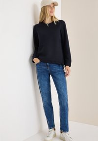 Marineblauw textuur sweatshirt met lange mouwen, gecombineerd met lichtblauwe jeans met zijsstrips en witte sneakers. Model staat tegen een neutrale achtergrond.