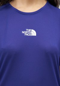 Άτομο που φοράει μπλε μπλουζάκι North Face με το λευκό λογότυπο της εταιρείας στο κέντρο του στήθους.