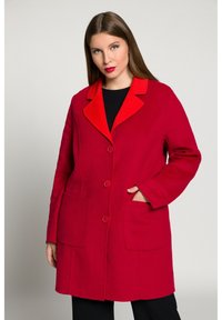 Cappotto in lana rossa con colletto arancione a contrasto, chiusura con bottoni e due tasche frontali. Presenta un design strutturato e dritto.