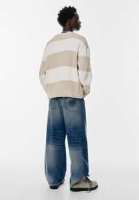 Gestreepte trui in beige en wit, oversized pasvorm, met ribbelstructuur. Gecombineerd met losse blauwe spijkerbroek en grijze schoenen.