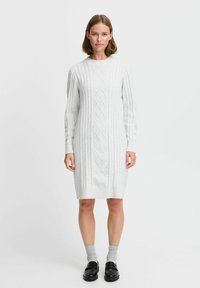 Hellgraues Strickkleid mit einem Kabelmuster, langen Ärmeln und Rundhalsausschnitt. Getragen mit grauen Socken und schwarzen Schuhen.
