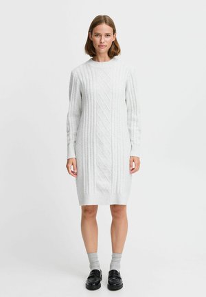 Lichtgrijze gebreide sweaterjurk met een kabelpatroon, lange mouwen en een ronde halslijn. Draagt met grijze sokken en zwarte schoenen.