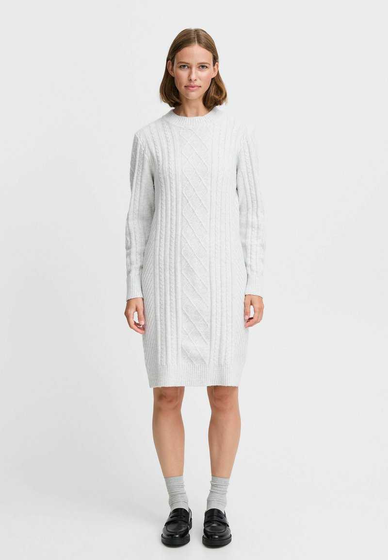 Hellgraues Strickkleid mit einem Kabelmuster, langen Ärmeln und Rundhalsausschnitt. Getragen mit grauen Socken und schwarzen Schuhen.